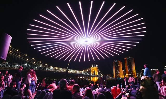 People gather to view a drone light show with the Roebling Suspension Bridge and buildings in the background.