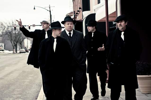 A group of men walk towards the camera dressed in stereotypical mob attire.