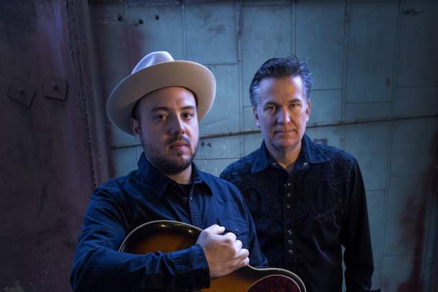 Two men look up at the camera. Both are in black. The one in the front holds a guitar and wears a hat.