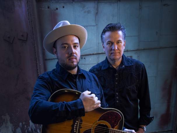 Two men look up at the camera. Both are in black. The one in the front holds a guitar and wears a hat.
