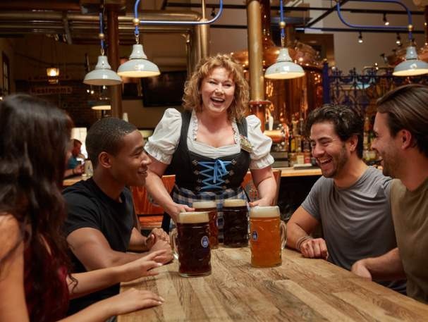A woman in traditional German dress serving four large beers to a group at Hofbrauhaus Newport