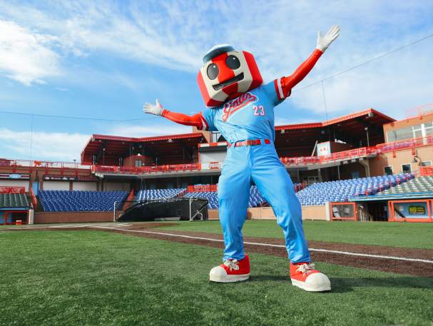 A red and white striped water tower mascot in a blue uniform stretches its arms wide with stadium seating behind.