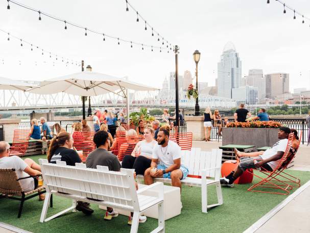 A group of people enjoy outside at Bridgeview Box Park. The Cincy skyline is in the background.