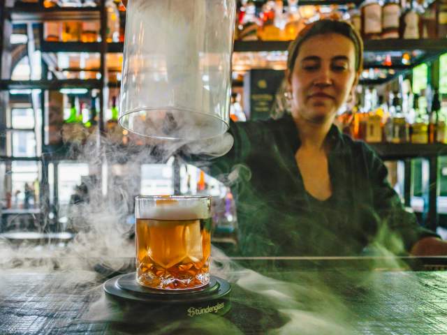 A bartender lifts the dome off a cocktail while smoke swirls around the bar.