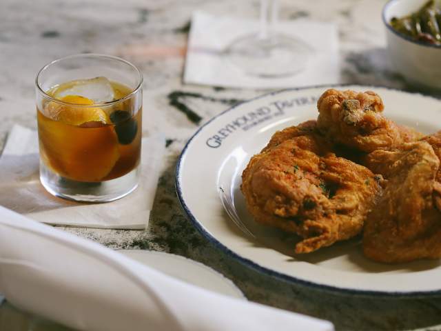A plate of fried chicken sits on a marbled table next to a cocktail and silverware roll.