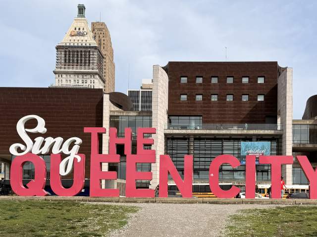 A large red and white statue spelling "Sing The Queen City" sits in a grassy park in front of a modern building and a tall skyscraper.