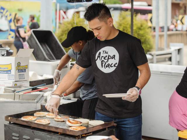 A person wearing a black shirt that says 'The Arepa Place" tends to food on a griddle at Taste of Cincinnati.