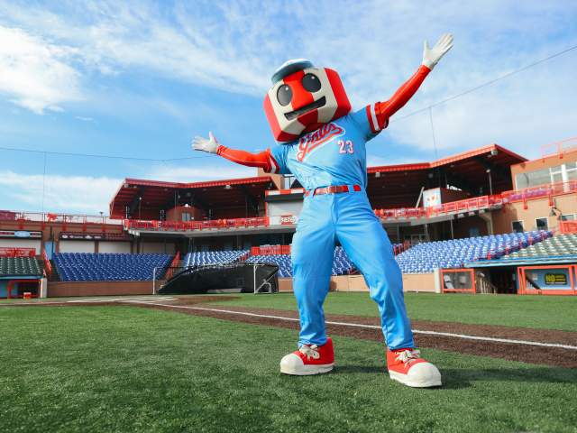 A red and white striped water tower mascot in a blue uniform stretches its arms wide with stadium seating behind.