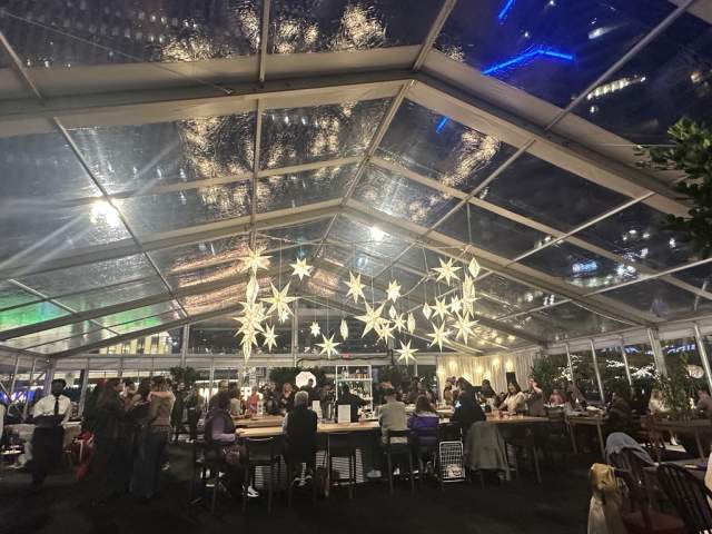 People are gathered in a tent with clear walls and roof. Glowing snowflakes hang from the ceiling.