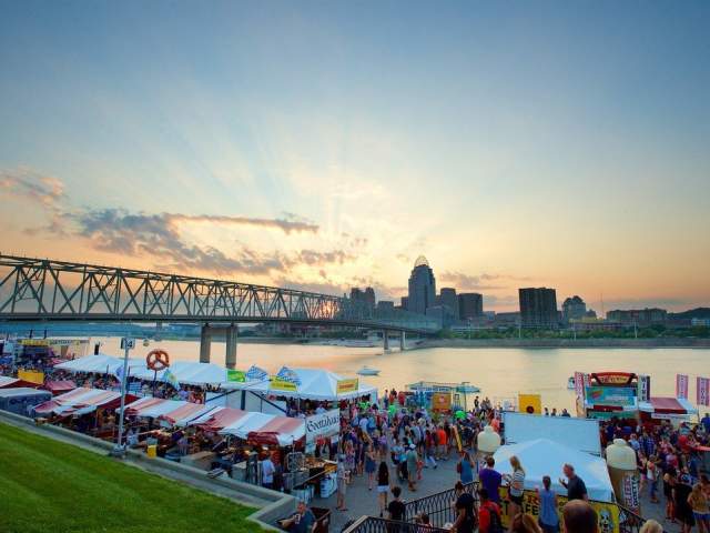 Image is of the Newport Festival Park with people walking around and the skyline of Cincinnati in the background.
