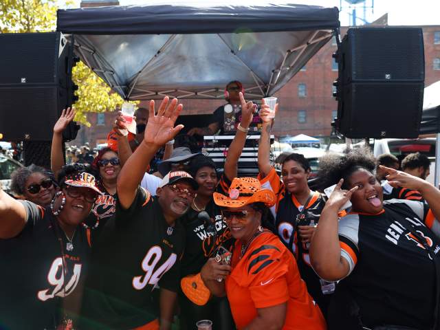A group of excited fans in Bengals jerseys cheer and wave in the foreground, while a DJ plays music under a canopy in the background.