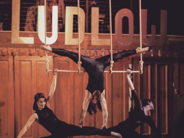 Image is of 3 trapeze artists in motion with the sign "Ludlow" behind them.