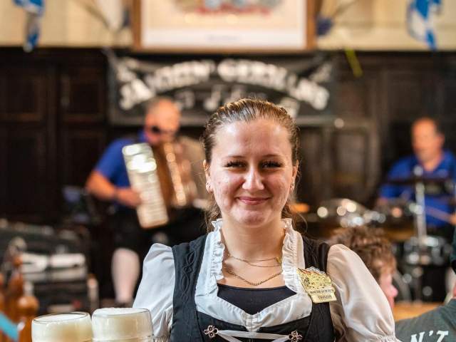 A smiling server walks towards the camera carrying two large beer steins in one hand.