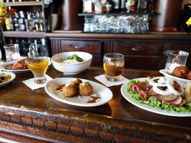 A wooden bar displays a spread of appetizers and drinks in the foreground, with a well-stocked bar and shelves of bottles in the background.