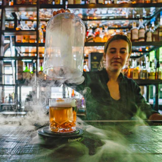 A bartender lifts the dome off a cocktail while smoke swirls around the bar.