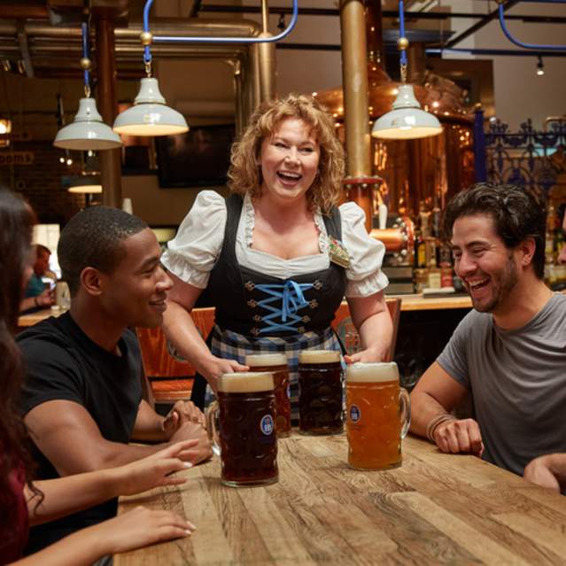 A woman in traditional German dress serving four large beers to a group at Hofbrauhaus Newport