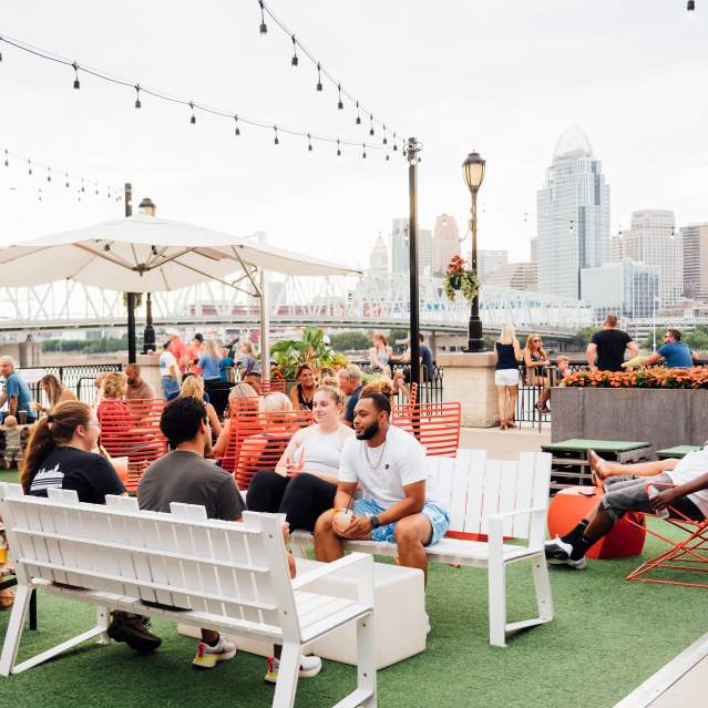 A group of people enjoy outside at Bridgeview Box Park. The Cincy skyline is in the background.