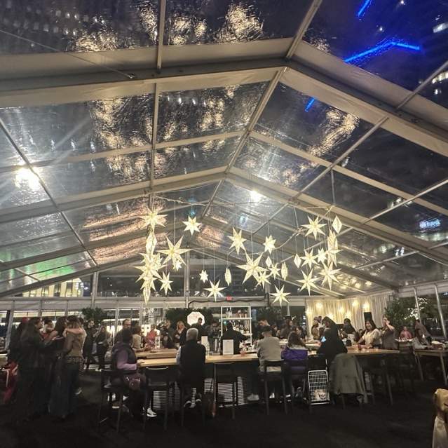 People are gathered in a tent with clear walls and roof. Glowing snowflakes hang from the ceiling.