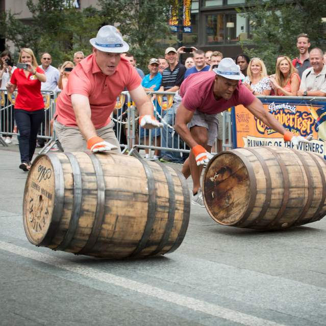 Two people in hats race down a street pushing barrels, while a crowd watches from behind a barricade in the background at Oktoberfest Zinzinnati.