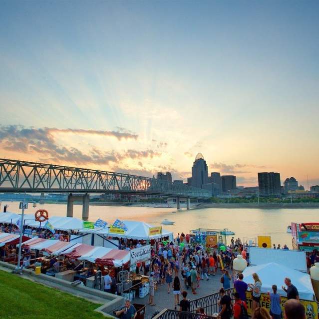 Image is of the Newport Festival Park with people walking around and the skyline of Cincinnati in the background.