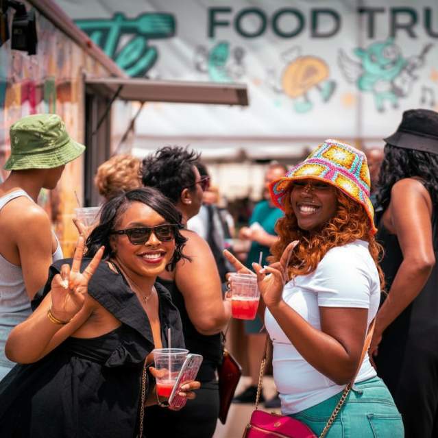 Two smiling women in the foreground hold drinks and flash peace signs in front of a colorful food truck with cartoon characters on its side.