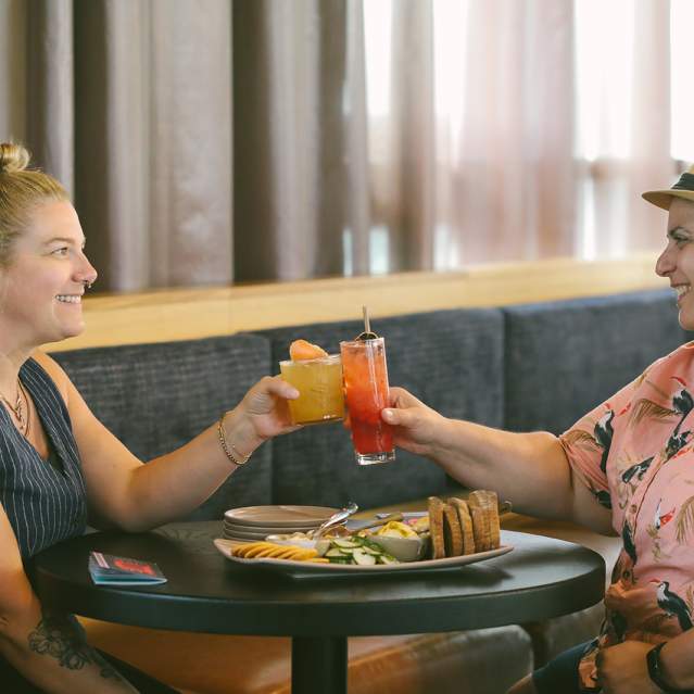 Two women toast each other with cocktails at a round table at the Aquifer bar at New Riff Distilling