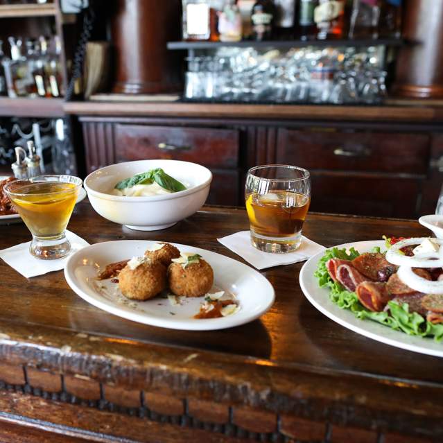 A wooden bar displays a spread of appetizers and drinks in the foreground, with a well-stocked bar and shelves of bottles in the background.