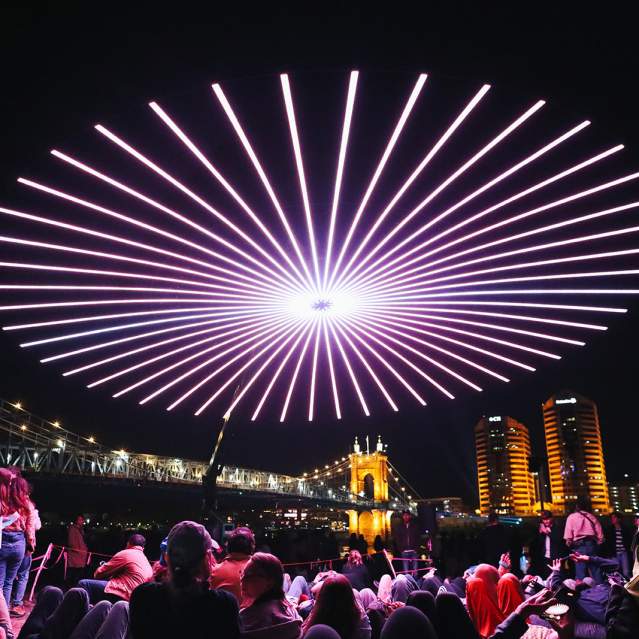People gather to view a drone light show with the Roebling Suspension Bridge and buildings in the background.
