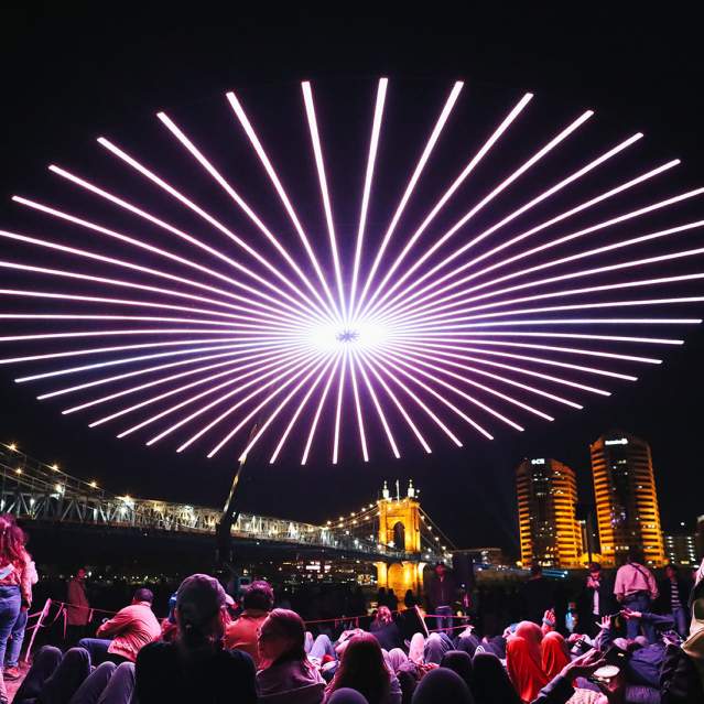 People gather to view a drone light show with the Roebling Suspension Bridge and buildings in the background.