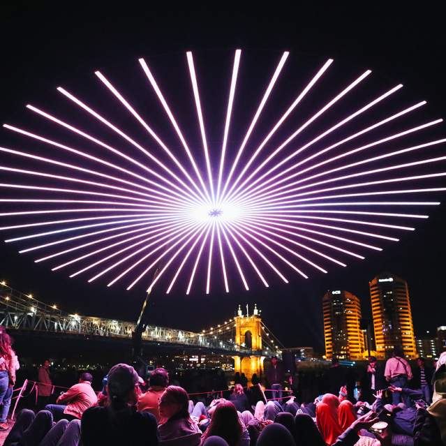 People gather to view a drone light show with the Roebling Suspension Bridge and buildings in the background.