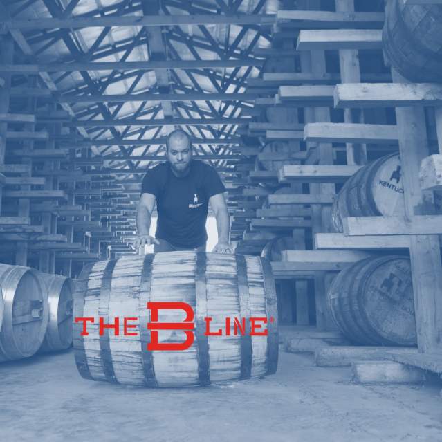 A man stands in the foreground, rolling a barrel while in the background, rows of barrels are stacked high on wooden shelves within a rickhouse. Over the photo says "The B-Line"