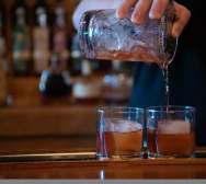 A close up of two drinks being poured over large, square ice in rocks glasses.
