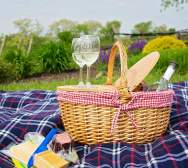 A picnic basket filled with wine and glasses sits on a checkered blanket in the foreground, with a lush green landscape and purple flowers in the background.