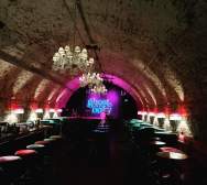 A tunnel lit with white and pink lights with a sign at the end in blue reading ghost baby. Rows of barstools line the walls of the tunnel