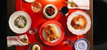 A red table is set with two place settings. One has a pasta with red sauce, the other a pasta with pesto and cheese. A bowl of olives and a plate of bread are between the two settings.