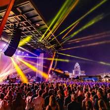 Night concert outdoors with rays of yellow light at Promowest Pavilion in Newport, Ky, with the Cincinnati skyline in the background