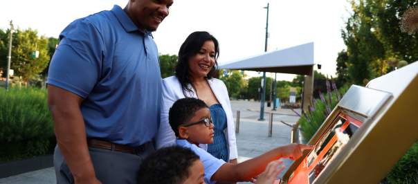 A family interacts with an outdoor display at the Black Music Walk of Fame in the foreground, while a park with trees and a covered structure is visible in the background.