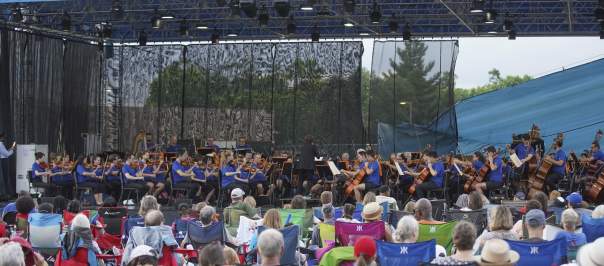 A crowd of people in pop-up lawn chairs sit by a stage and watch the Cincinnati Symphony Orchestra perform