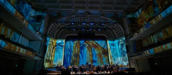 The Cincinnati Symphony Orchestra plays in front of a lit up back drop.
