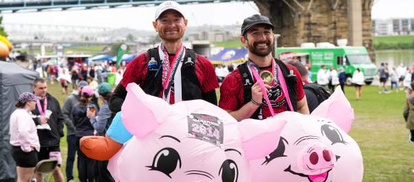 Two men after Flying Pig with medals in flying pig costumes in front of the Roebling Bridge