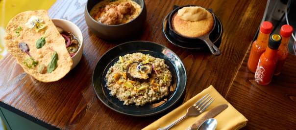 A yellow napkin with silverware sits in front of several plates on a table including a rice dish, cornbread, and a stew with hot sauce bottles to the right.