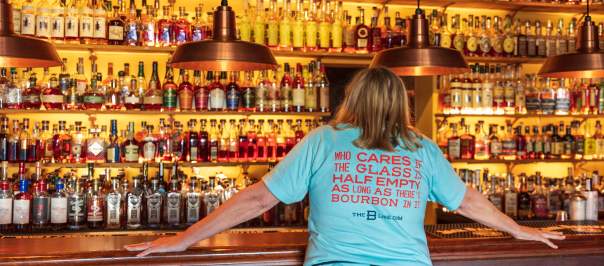 A woman in a light blue shirt stands in front of seemingly hundrands of bourbon botttles. Her shirt reads, "Who cares if the glass is half empty as long as there's bourbon in it" The B-Line