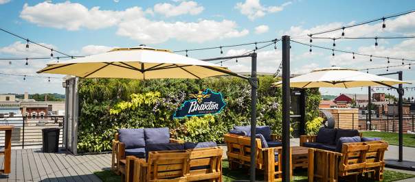 A patio set, umbrellas and string lights sits on a rooftop deck in front of a living plant wall that says Braxton