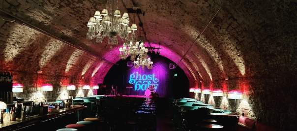 A tunnel lit with white and pink lights with a sign at the end in blue reading ghost baby. Rows of barstools line the walls of the tunnel