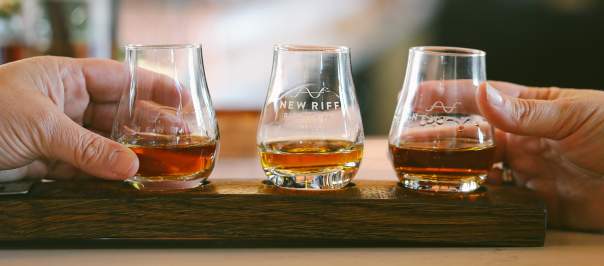 Three glencairn glasses, each with a pour of whiskey, sit in a wooden holder. Hands from either side reach out to select a glass.