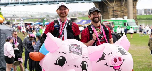 Two men after Flying Pig with medals in flying pig costumes in front of the Roebling Bridge
