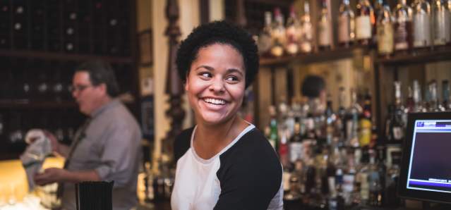 Young Black woman smiling with the bourbon collection of Tousey House Tavern behind her