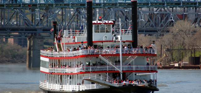 A red and white, paddlewheel riverboat cruises on the water. There are 3 decks with people standing on them.