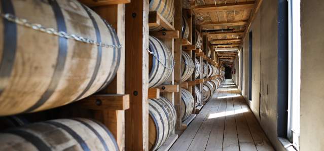 Rows of bourbon barrels line the left side of the photo in a wooden rickhouse.