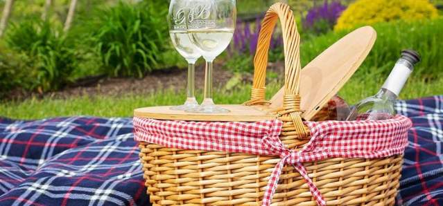 A picnic basket filled with wine and glasses sits on a checkered blanket in the foreground, with a lush green landscape and purple flowers in the background.